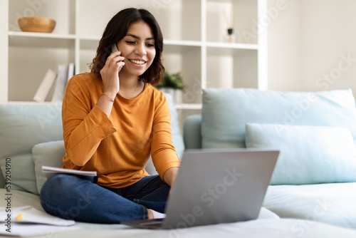 Remote Business. Young Middle Eastern Woman Using Cellphone And Laptop At Home