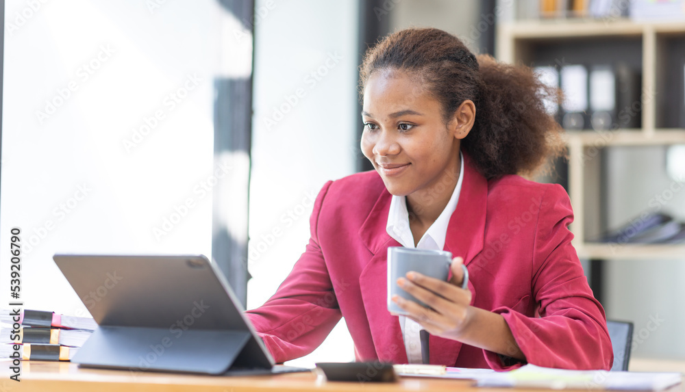 Smiling young African american female Finance Entrepreneur sitting in workplace office hoding coffee cup and working on a laptop, investment financial document report finance analysis concept. Smiling young African american female Finance Entrepreneur sitting in workplace office hoding coffee cup and working on a laptop, investment financial document report finance analysis concept.