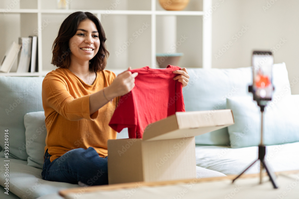 Smiling Arab Female Influencer Unpacking Clothes While Recording Video Content At Home Smiling Arab Female Influencer Unpacking Clothes While Recording Video Content At Home