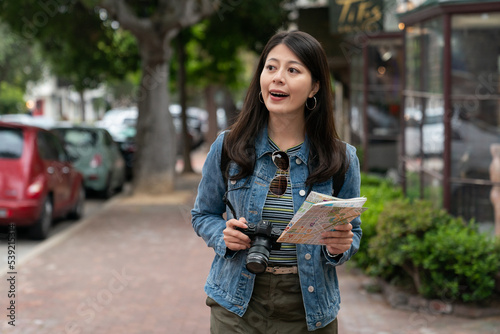 happy asian female tourist having fun exploring the charming beach town Carmel by the sea with camera and city map. she looks around her surroundings while taking stroll on street © PR Image Factory