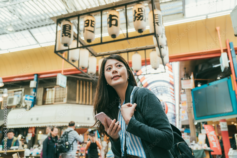 cheerful asian girl using cellphone for online information and looking around enjoying active vibe in kuromon ichiba market in Osaka japan. translation:â kuromon ichiba marketâ cheerful asian girl using cellphone for online information and looking around enjoying active vibe in kuromon ichiba market in Osaka japan. translation:â kuromon ichiba marketâ