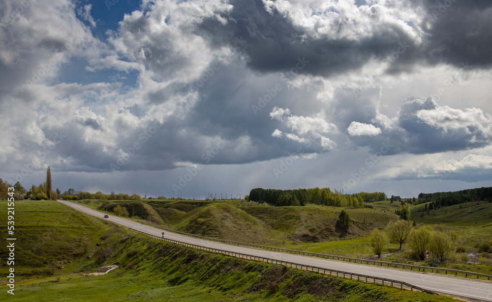 Osphalt road. Spring landscape, dramatic sky over green hills, before rain. The sun through the clouds illuminates the road and ravines.