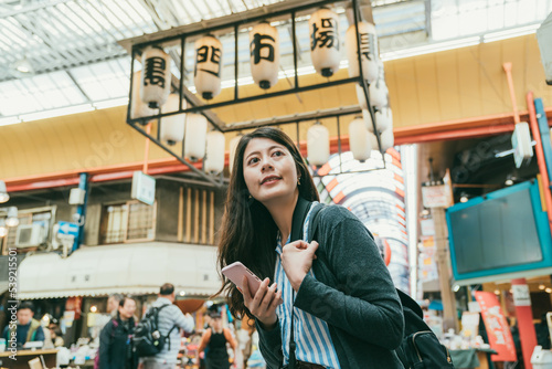 cheerful asian girl using cellphone for online information and looking around enjoying active vibe in kuromon ichiba market in Osaka japan. translation:â kuromon ichiba marketâ photo