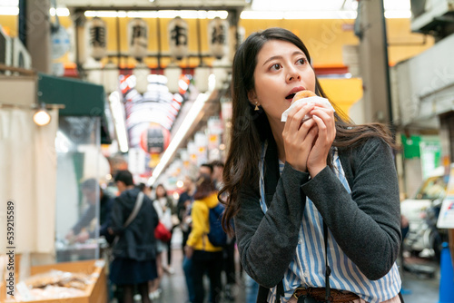 Taiwanese woman tourist taking a bite into donut and looking around feeling curious about various food stalls in kuromon ichiba market in Osaka japan photo