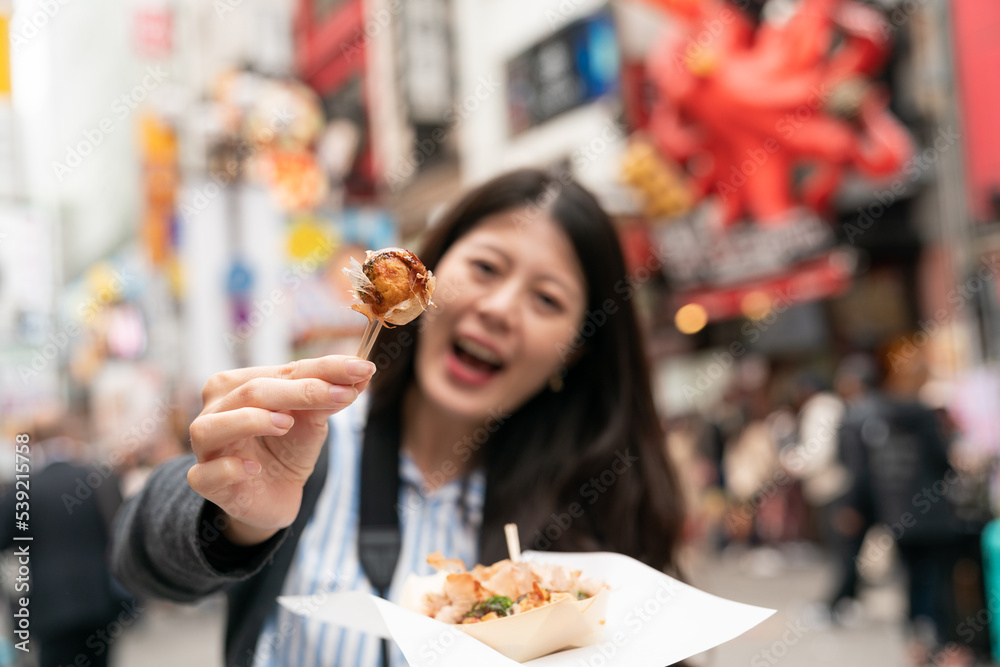 selective focus cheerful Asian girl tourist facing camera and saying ah while feeding boyfriend takoyaki ball on the shopping street in shinsaibashi suji and doutonbori Osaka japan selective focus cheerful Asian girl tourist facing camera and saying ah while feeding boyfriend takoyaki ball on the shopping street in shinsaibashi suji and doutonbori Osaka japan