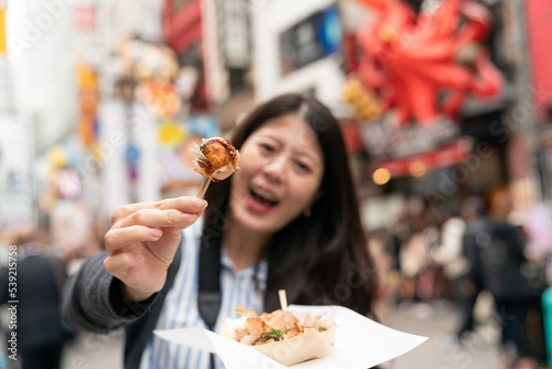 selective focus cheerful Asian girl tourist facing camera and saying ah while feeding boyfriend takoyaki ball on the shopping street in shinsaibashi suji and doutonbori Osaka japan