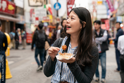 cheerful Asian woman traveler having fun looking at various Japanese restaurants while eating takoyaki and walking in shinsaibashi suji and doutonbori area in Osaka japan