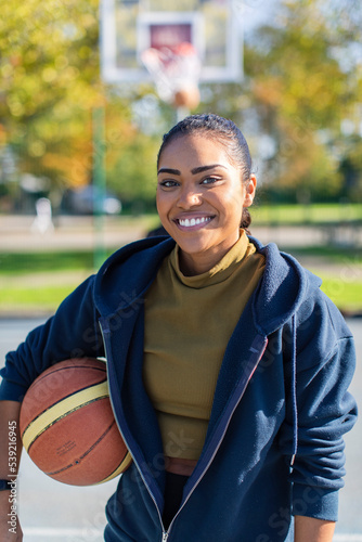 Portrait of young female holding a basketball and smiling to camera as she stands on a basketball court outdoors
