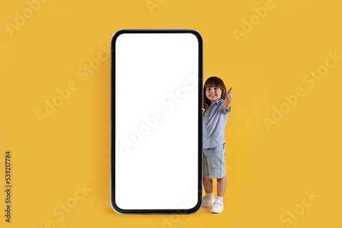 . Adorable little boy standing near big smartphone with blank white screen and showing thumb up gesture, empty space