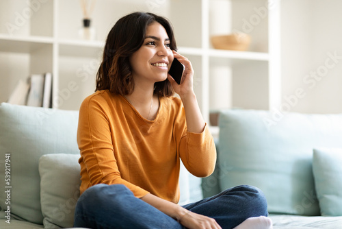 Young Arab Woman Talking On Cellphone And Relaxing On Couch At Home