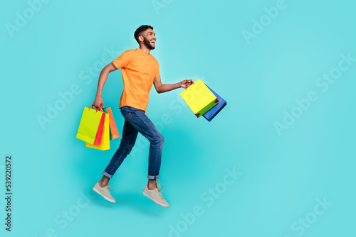 Full length profile photo of energetic guy hold packages jump run hurry empty space isolated on teal color background