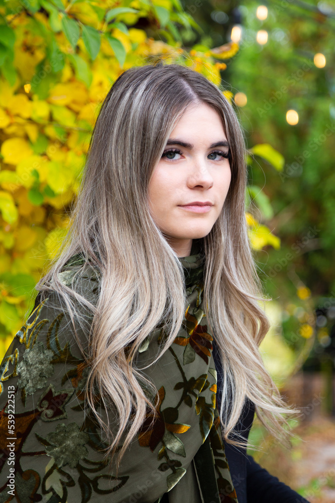 Naklejka premium Vertical portrait of beautiful blue-eyed young woman with long dirty blond hair standing outdoors with fall foliage in soft focus background