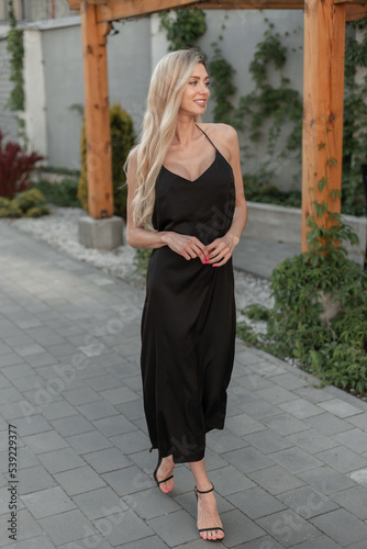 Fashionable happy pretty woman model with blonde hair in black stylish long strappy dress with shoes walks on the street