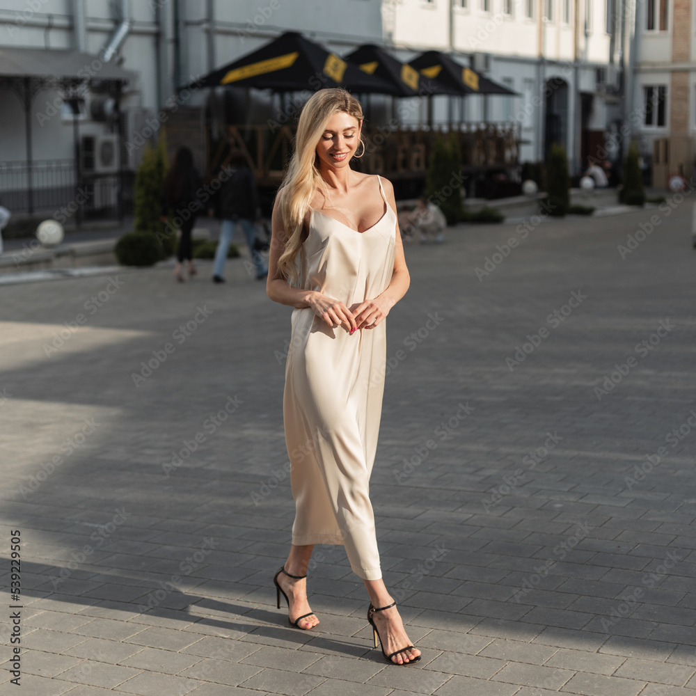 Happy beautiful young woman with a smile in a fashionable beige dress with strappy heels shoes walking in the city in the sunshine