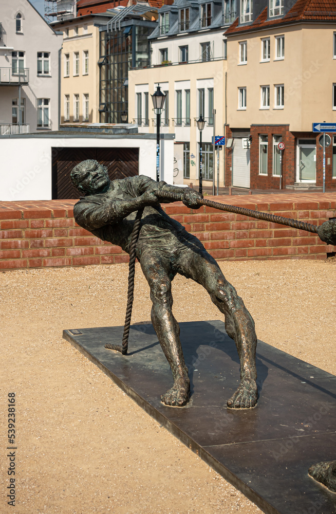 Naklejka premium Germany, Wismar - July 13, 2022: St. Marienkirche. Detail of 1 man at Tauziehen or tug-of-war statue in front. Multicolor house facades around square in back. Yellow gravel bottom