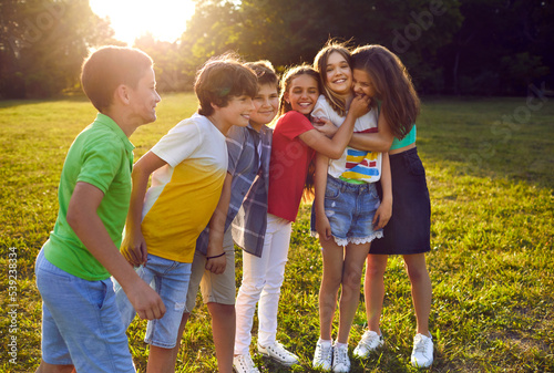 Friendly funny teen boys and girls in summer casual clothes have good time outdoors together with peers or school buddies and hug each other and play in sunny green park. Active kids rest