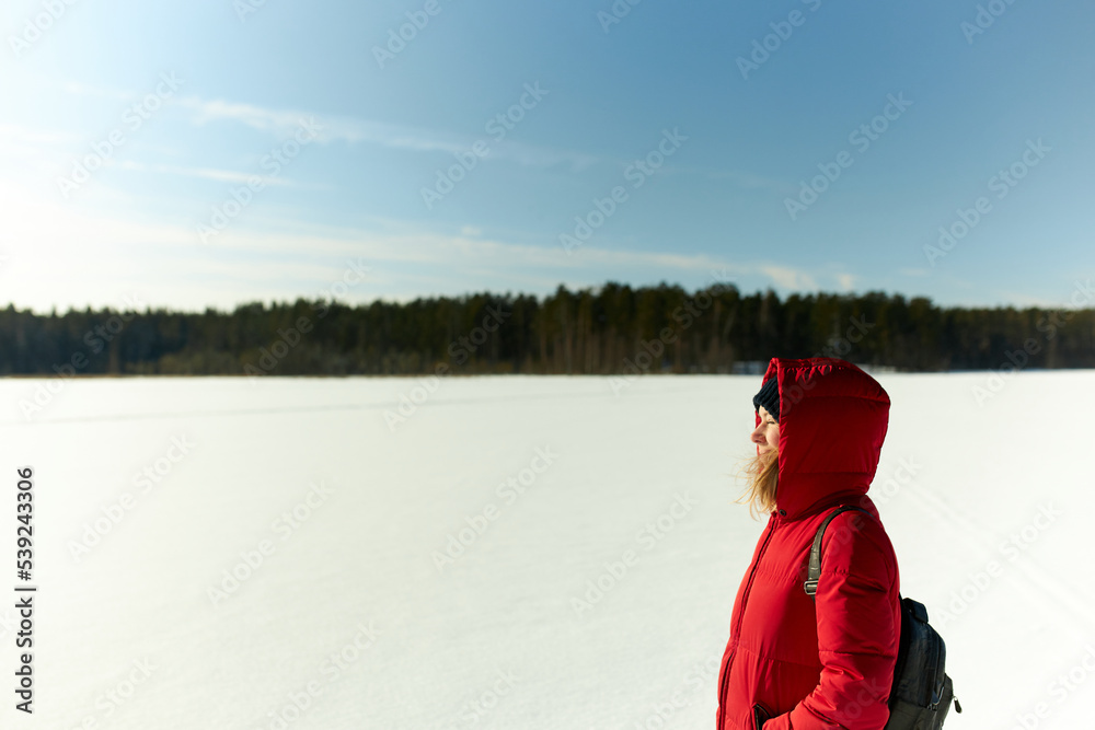Side view of female in warm red puffer carrying backpack on shoulders enjoying fresh air and tranquility of wild nature, keeping eyes closed on bright sun and sparkling snow, copy space on the right
