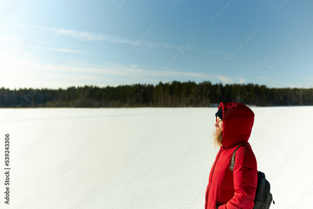 Side view of female in warm red puffer carrying backpack on shoulders enjoying fresh air and tranquility of wild nature, keeping eyes closed on bright sun and sparkling snow, copy space on the right Side view of female in warm red puffer carrying backpack on shoulders enjoying fresh air and tranquility of wild nature, keeping eyes closed on bright sun and sparkling snow, copy space on the right