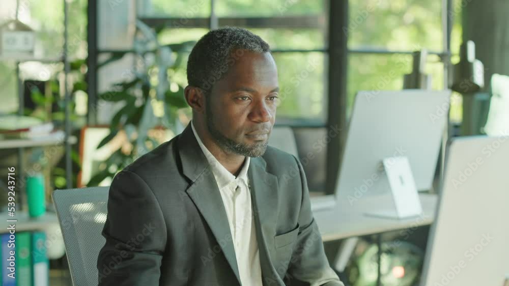 Portrait african american man turns his head to look at the camera smiling using computer. Sitting in office. Businessman, entrepreneur, CEO. Corporate business. Slow motion Portrait african american man turns his head to look at the camera smiling using computer. Sitting in office. Businessman, entrepreneur, CEO. Corporate business. Slow motion