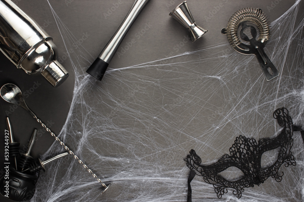 Various bar accessories such as: a shaker, muddler, strainer, as well as a bar spoon and a measuring stack lie against the backdrop of a spider web on a black matte table. Adult halloween celebration Various bar accessories such as: a shaker, muddler, strainer, as well as a bar spoon and a measuring stack lie against the backdrop of a spider web on a black matte table. Adult halloween celebration
