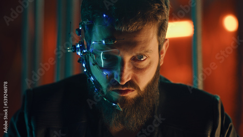 Young bearded guy in a black leather jacket looks fiercely at the camera. Guy with one-eyed glasses and headset trapped behind black thick rods hanging to prevent him from escaping. © Framestock