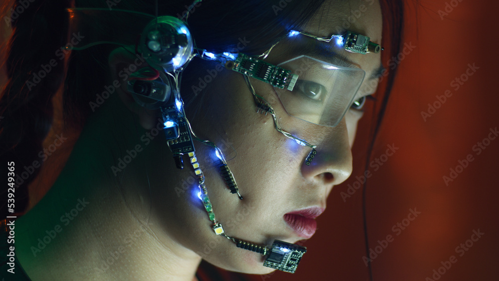 Focused asian girl in cyberpunk attire works on the computer. Wearing one-eyed glasses with white LED lights and a microphone. Neon lights background. science fiction, cybernetics concept. Focused asian girl in cyberpunk attire works on the computer. Wearing one-eyed glasses with white LED lights and a microphone. Neon lights background. science fiction, cybernetics concept.