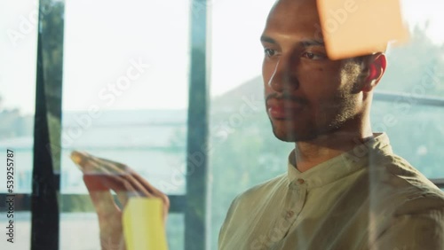 Close-up portrait of thoughtful young African man standing near glass board and looking at colorful sticky notes. Good-looking successful intelligent guy planning project in office. Indoors