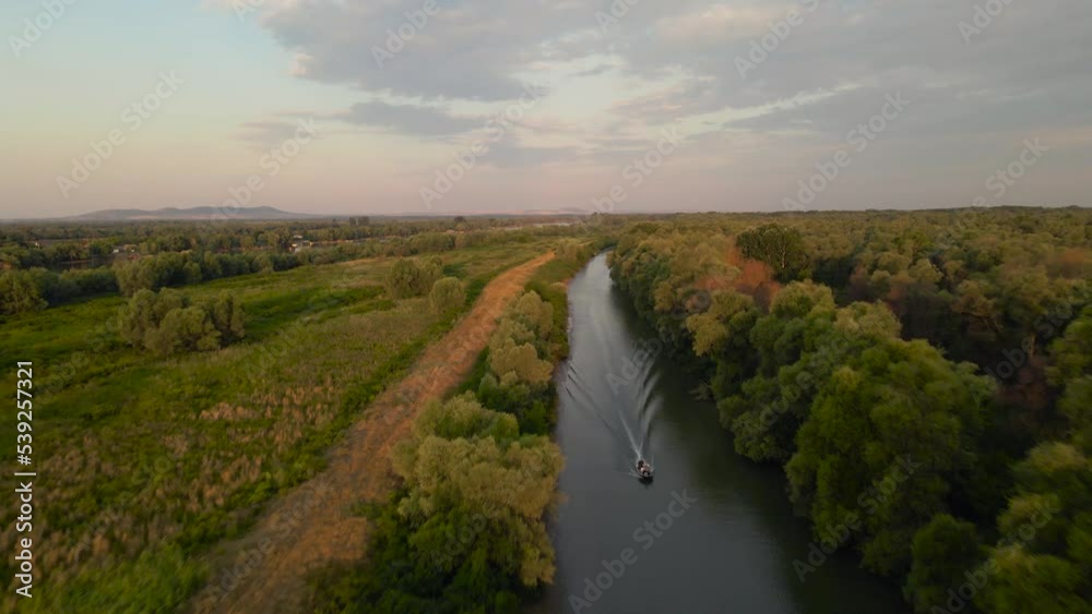 Aerial footage of a canal in Danube's delta, shot from a drone while flying backwards, with a motorboat and beautiful sunrise scenery.
