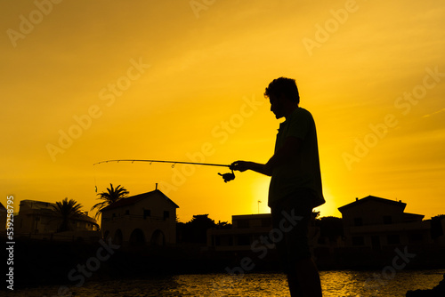 Angler in silhouette looking at the fishing rod before throwing it into the sea, behind a coastal town and the sea, orange sunset background