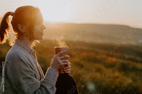 Woman enjoying hot tea from thermo cup at sunset on a hill with rural background photo