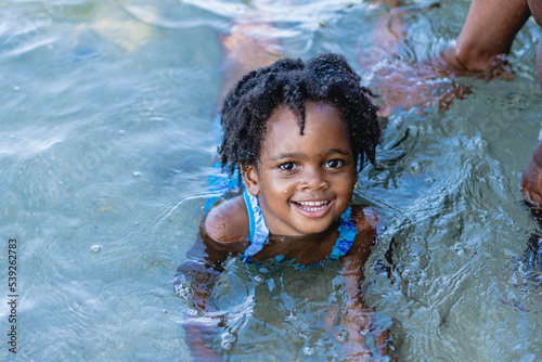 Retrato horizontal de una pequeña niña afroamericana de cabello afro muy sonriente y mirando a cámara acostada boca abajo a la orilla del mar jugando.  photo