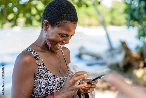 Imagen horizontal de una hermosa mujer africana de cabello corto muy sonriente sola en la playa utilizando un celular.  photo
