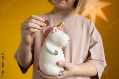 The girl holds a unicorn piggy bank in her hands and puts a coin in it.