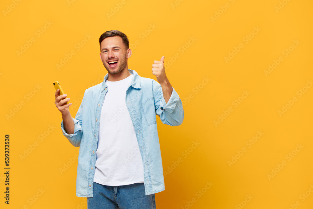 LUCKY WIN OFFER. Happy tanned handsome man in blue basic t-shirt raise thumb up hold phone posing isolated on orange yellow studio background. Copy space Banner Mockup. Online Social media concept LUCKY WIN OFFER. Happy tanned handsome man in blue basic t-shirt raise thumb up hold phone posing isolated on orange yellow studio background. Copy space Banner Mockup. Online Social media concept