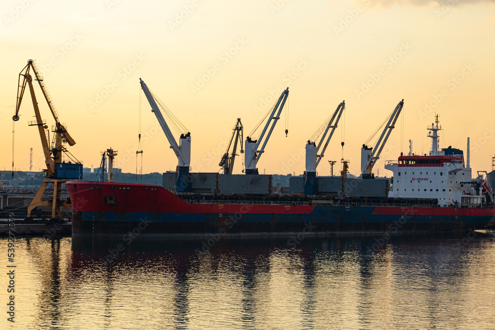 Sea port in Constanta, Romania at sunset with a moored cargo ship at the pier and port cranes on site.