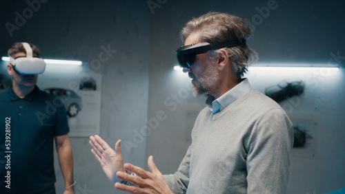 Two automotive professional engineers wearing virtual reality glasses talk about vehicle production while standing in a high tech development laboratory. Car design analysis and improvement.