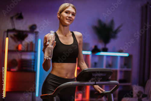 Portrait of active and dynamic young charming thirsty woman holding water bottle doing sport fitness at home running on treadmill indoor at night. Tone your body. Perfect shape. Improving endurance.