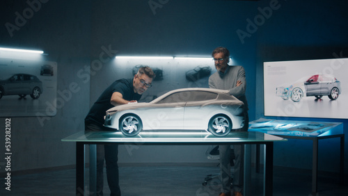 Experienced automotive engineers discuss the design infront of a rake sculpture prototype car model. Engineers use a touch screen digital tablet in a high tech laboratory. Make design corrections.