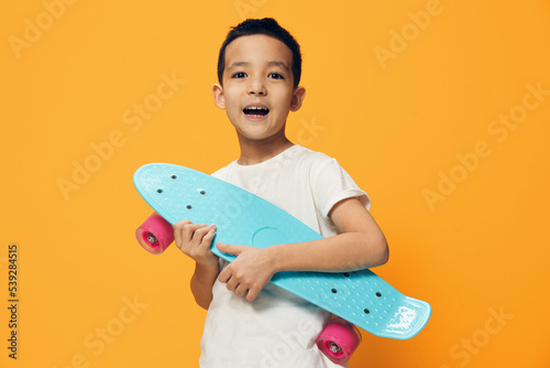 a very joyful attractive boy of school age stands on an orange background smiling broadly at the camera and holding a blue small skateboard in his hand