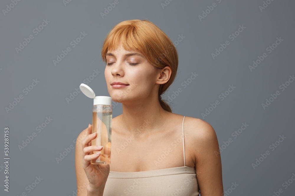 Close-up photo.Horizontal studio shot. An emotional woman in a white T-shirt with light, clear skin, with shiny red hair gathered in a ponytail on a gray background with a transparent bottle of water Close-up photo.Horizontal studio shot. An emotional woman in a white T-shirt with light, clear skin, with shiny red hair gathered in a ponytail on a gray background with a transparent bottle of water