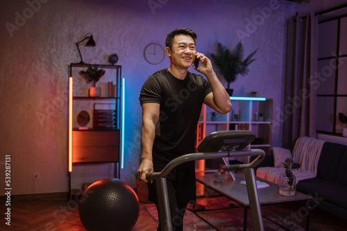 Portrait of young asian sportive man in dark sportswear talking on smartphone while training at home, doing cardio exercise on treadmill at evening time. Concept of sport, health care, communication.