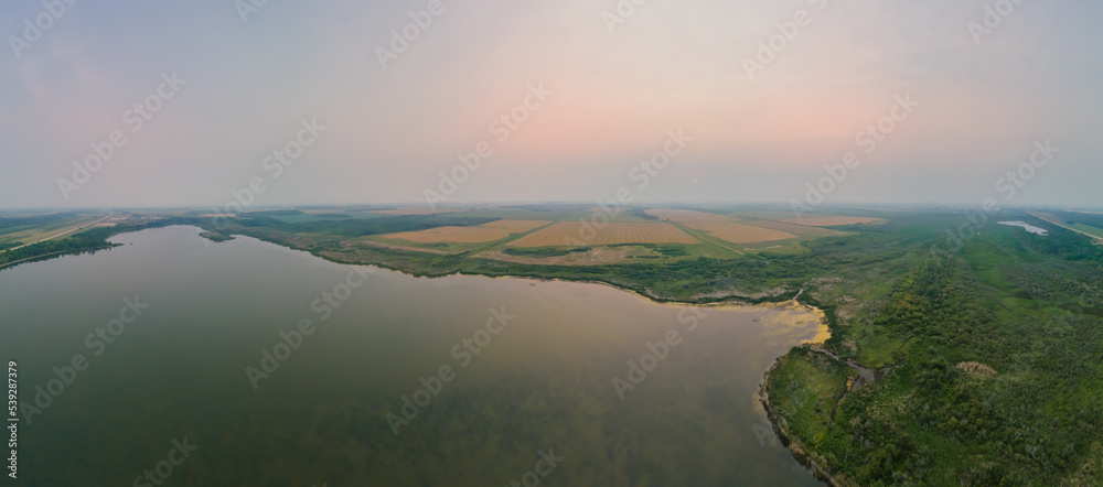 Foggy morning aerial looking over a large sallow lake with forest and farm fields in the distance.
