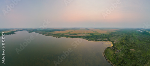 Foggy morning aerial looking over a large sallow lake with forest and farm fields in the distance. 