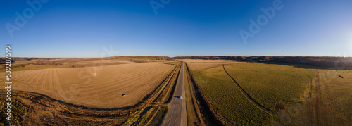 Aerial view of a two-lane paved highway crossing large farm fields with distant trees and a clear blue sky 