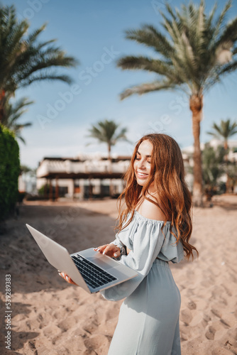 A cheerful blogger girl remotely works with a laptop on the beach with palm trees. A woman makes a video from a trip. Travels the world and works with social networks online. Remote modern profession © Daria Lukoiko