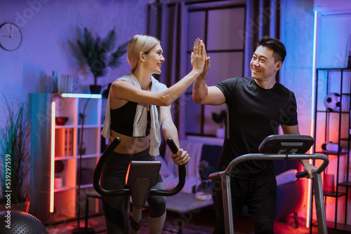 Joyful caucasian woman training on exercise bike with sporty asian man running on treadmill and giving high five. Smiling young couple doing cardio training on exercise machines at evening time