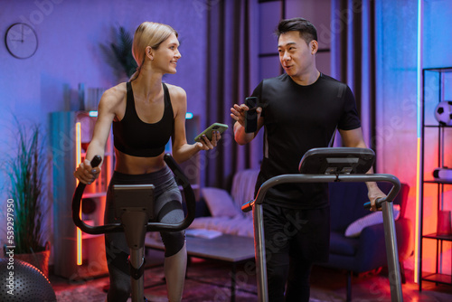 Fototapeta Naklejka Na Ścianę i Meble -  Blond caucasian sports women in sportswear cycling bike and fit asian man running on treadmill and showing each other photos from social networks.