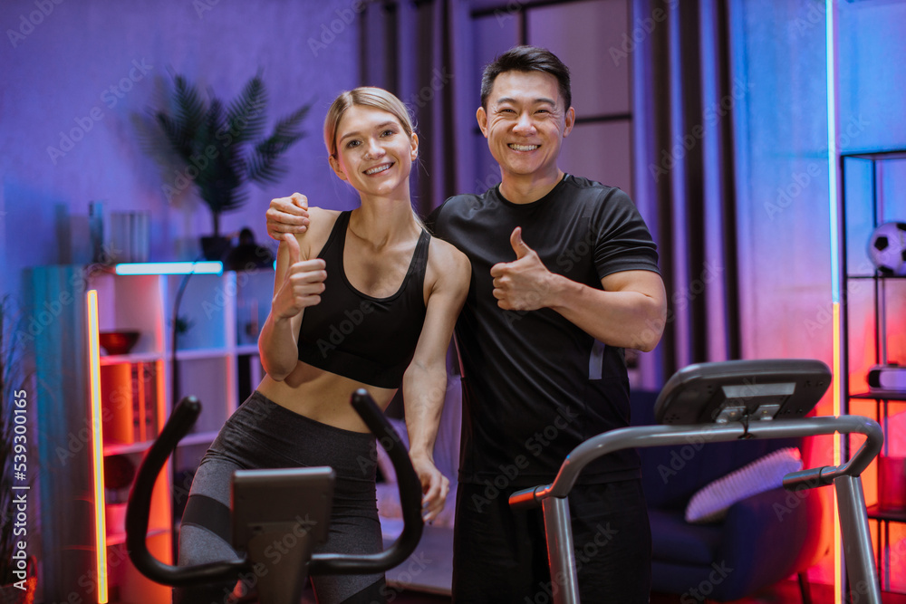 Embracing young couple doing cardio training on exercise machines showing thumbs up at evening time. Happy woman training on exercise bike with handsome sporty asian man running on treadmill. Embracing young couple doing cardio training on exercise machines showing thumbs up at evening time. Happy woman training on exercise bike with handsome sporty asian man running on treadmill.