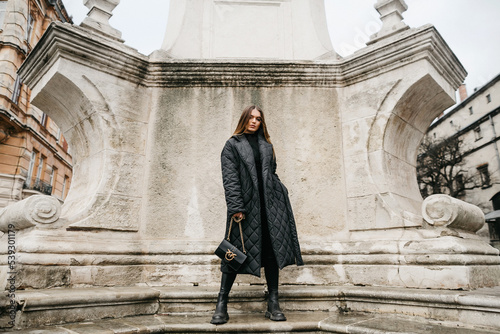 Front view of confident woman in stylish outfit posing near part of old building. Fashionable girl with brown long hair, dressed in quilted down jacket and holding bag, standing in ancient town