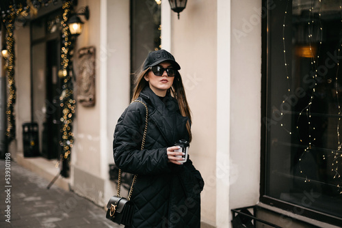 Side view of woman tourist in fashionable outfit walking in town. Girl in cap and sunglasses, wearing in trendy jacket, holding cup of coffee and enjoying atmosphere of Christmas in town